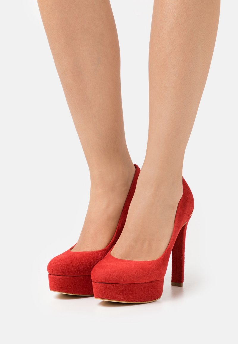 Jambes portant des escarpins compensés en daim rouge avec des talons aiguilles épais sur fond blanc.