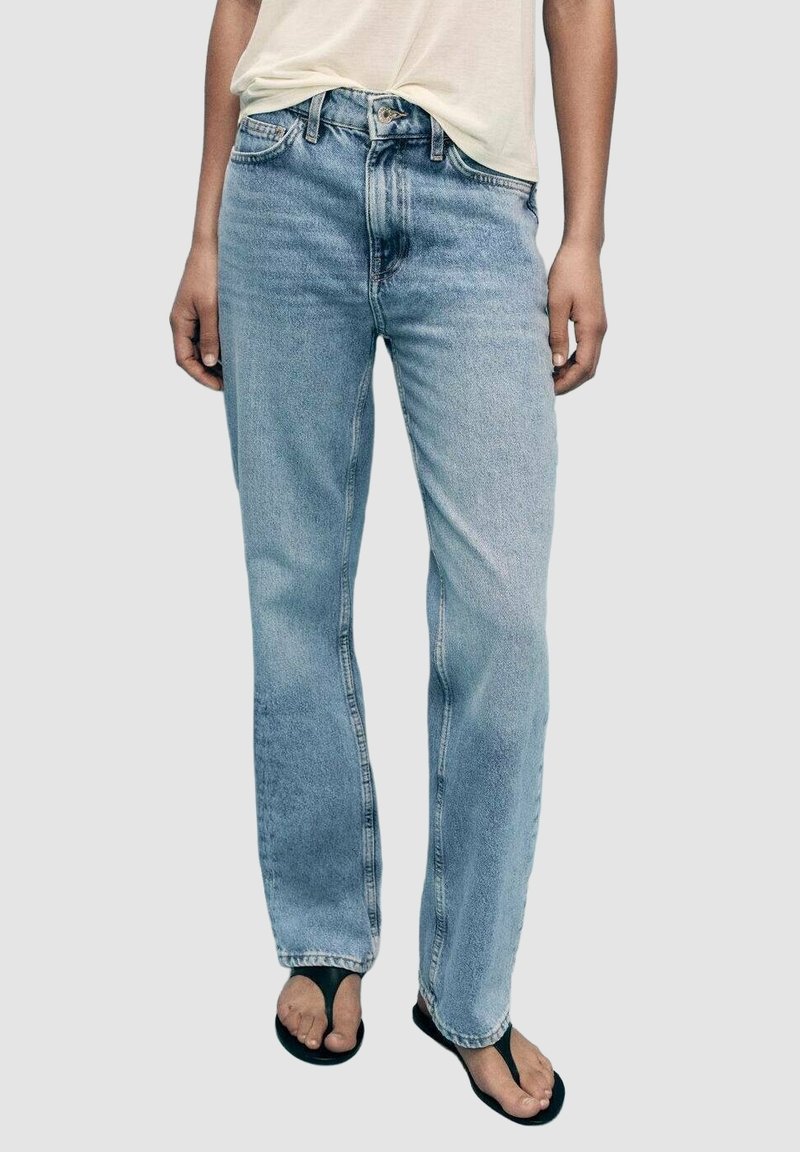 Personne portant un jean bleu clair à jambes droites, une chemise crème rentrée et des sandales noires à lanières sur un fond uni.
