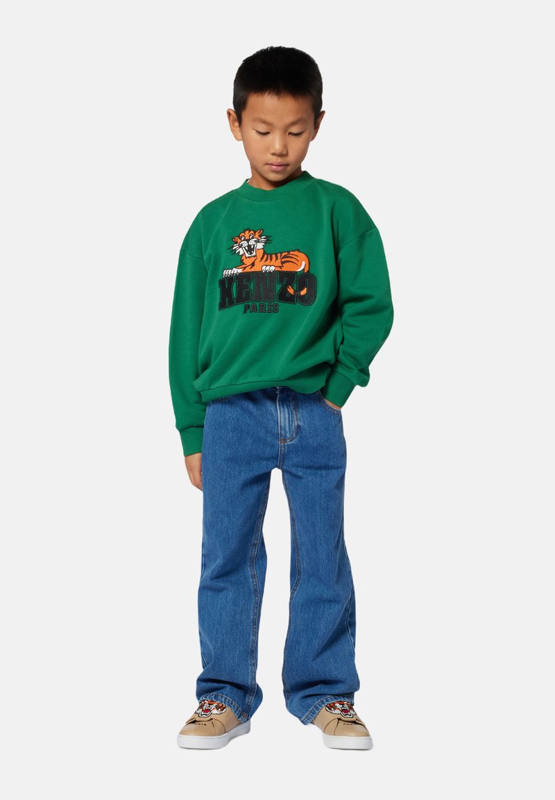 Zielona bluza z graficznym motywem tygrysa i napisem "KENZO PARIS", zestawiona z niebieskimi dżinsami i beżowymi sneakersami z motywem tygrysa.