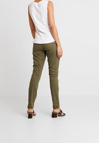 Olivgröna byxor med slim fit och en jämn textur, kombinerat med en ärmlös vit topp och svarta klacksandaler, sett bakifrån.