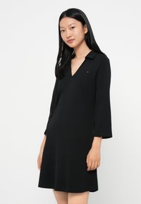 Czarna sukienka z dekoltem w kształcie litery V i rękawami trzyczwartkowymi. Wykonana z gładkiego materiału, z małym logo na piersi i rozkloszowanym dołem.