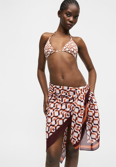 Γυναίκα που φοράει μπικίνι τοπ με καφέ και λευκό σχέδιο και αντίστοιχο σαρόνγκ δεμένο στη μέση, στέκεται μπροστά σε απλό φωτεινό φόντο.