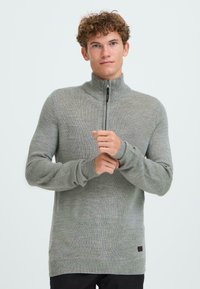 Grå zip-neck genser laget av et teksturert strikket stoff, med høy krage og ribbede mansjetter. Diskret merkevarelabel på den nedre siden.