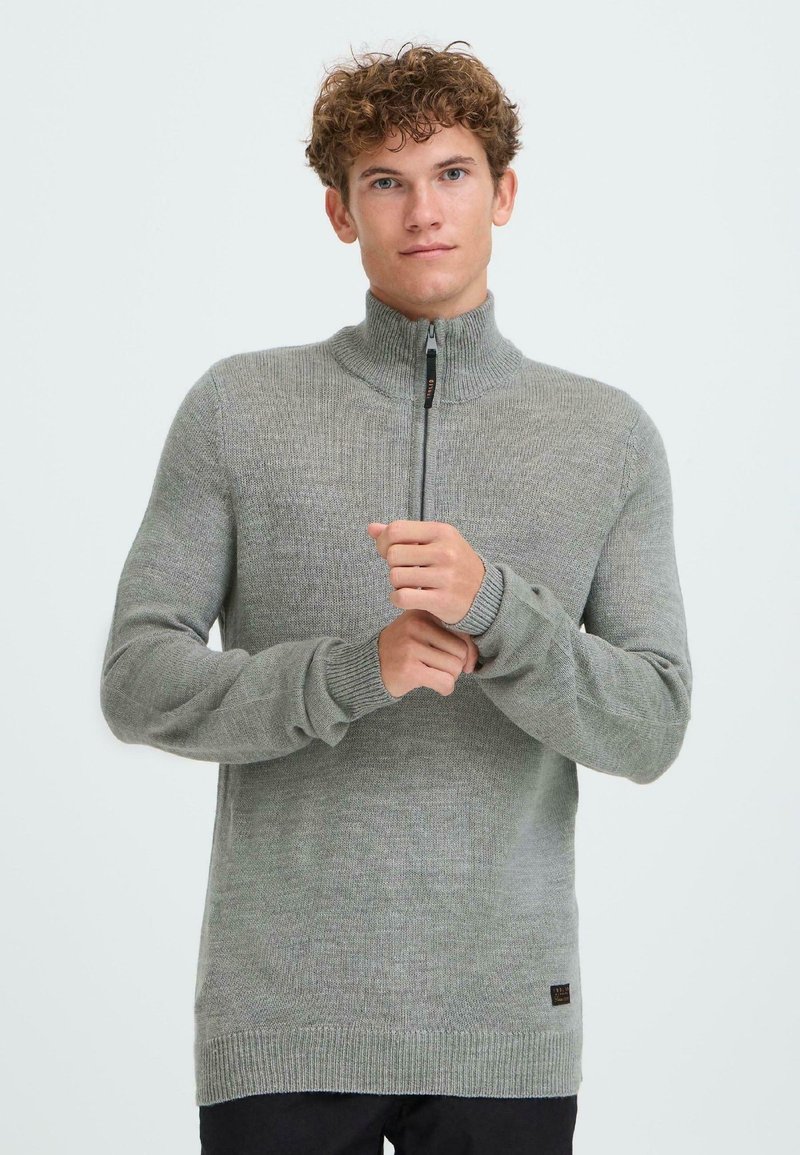 Grå zip-neck genser laget av et teksturert strikket stoff, med høy krage og ribbede mansjetter. Diskret merkevarelabel på den nedre siden.