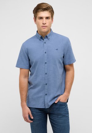 Junger Mann mit blauem kurzärmligem Hemd mit Knöpfen und dunkler Jeans, der mit einer Hand in der Tasche vor einem einfarbigen Hintergrund steht.