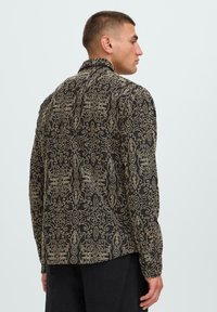 Črna dolga majica z dolgimi rokavi z zapletenimi zlatimi paisley in cvetličnimi vzorci, ki ima tradicionalen ovratnik in raven kroj.