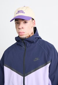 Veste zippée marine et violet clair avec capuche, dotée d'une bande horizontale noire et du logo Nike. Casquette beige avec texte violet.