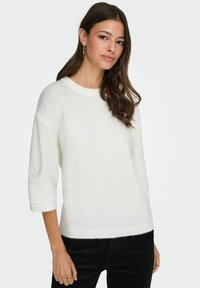 Pull blanc et moelleux avec un col rond et des manches trois-quarts, présentant des poignets et un ourlet côtelés. Associé à un jean foncé.