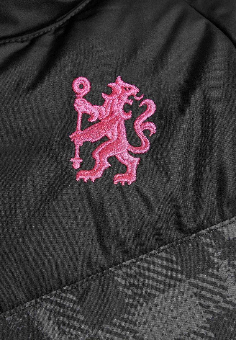 Tela negra con un patrón texturizado debajo de un prominente emblema de león bordado en rosa sosteniendo un bastón. Superficie suave, diseño elegante.