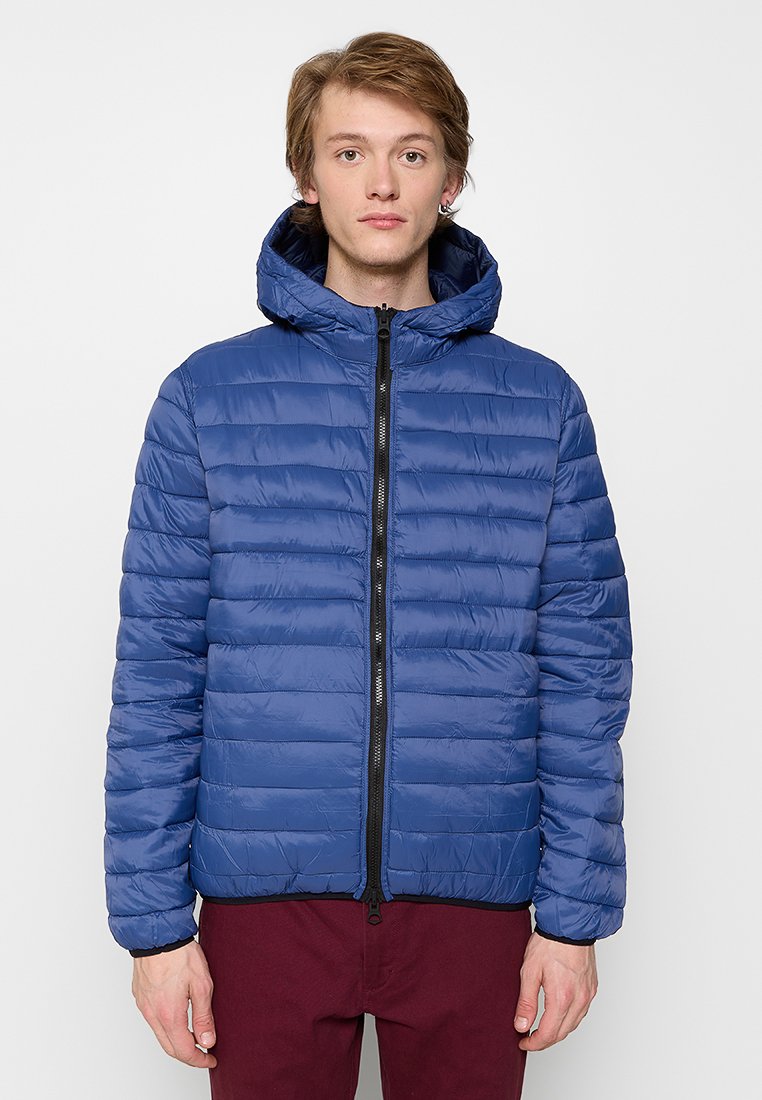 Joven con cabello corto, vistiendo una chaqueta acolchada con capucha azul y pantalones burdeos, de pie frente a un fondo claro y sencillo.