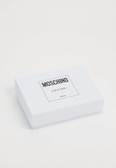 Valge ristkülikukujuline karp, millel on must silt "Moschino Couture!" musta tekstiga, mida ümbritseb õhuke must joon. Sujuv tekstuur, minimalistlik disain.