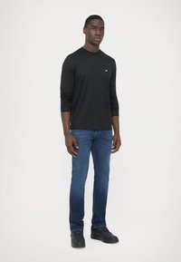 Μαύρο μακρυμάνικο πουκάμισο με στρογγυλή λαιμόκοψη, συνδυασμένο με μπλε τζιν παντελόνι και μαύρα αθλητικά παπούτσια. Απλός σχεδιασμός με ελάχιστες λεπτομέρειες.