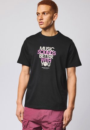 Ung mann med kort krøllete hår som har på seg svart t-skjorte med teksten "Music sounds better with you" og rosa cargoshorts, ser til siden.