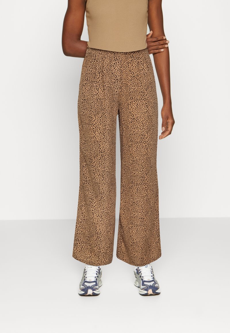 Calças largas castanhas com padrões e manchas pretas, feitas de um tecido suave. Usadas com uma blusa ajustada bege e ténis brancos.