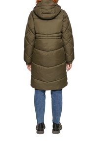 Olijfgroene pufferjas met een capuchon, voorzien van een doorgestikt patroon en ribbelmanchetten. Draagt over blauwe jeans en zwarte laarzen.