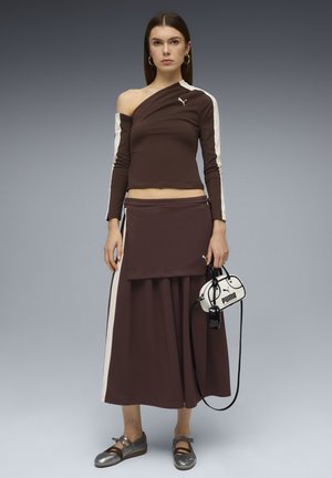 Brun langærmet top med asymmetrisk design og cremelignende striber på siderne, kombineret med et matchende skørt og skinnende sølvfarvede sko. Lille håndtaske.