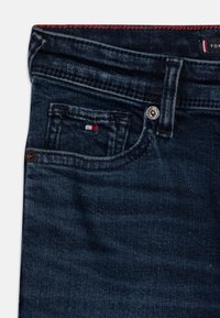 Granatowe dżinsy z przodu posiadające kieszeń z małym, trójkolorowym detalem logo, kontrastowe przeszycia oraz metalowy guzik zapięcia.