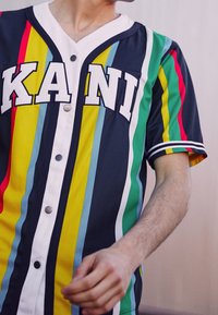 Baseballshirt met meerkleurige verticale strepen in marineblauw, geel, groen en rood. Heeft witte letters "KANI" en drukknopen.