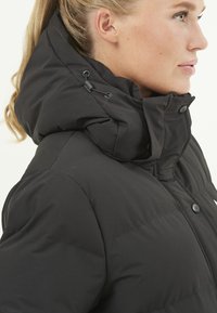 Veste matelassée noire avec un col haut, des cordons de serrage réglables et des boutons à l'avant. Texture lisse avec un ajustement sécurisé autour du cou.