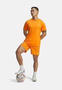 Vyrų futbolininkas oranžinėje aprangoje stovi su viena koja ant baltos ir raudonos futbolo kamuolio, priešais vientisą baltą fono.