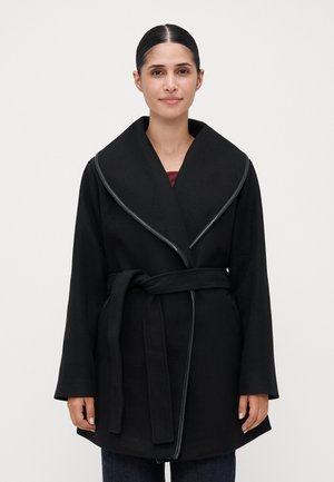 Woman with dark hair tied back wearing a black belted coat with wide collar over dark pants, standing against a plain light background.
