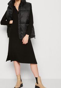 Mustat pehmustetut liivit pystykolarilla sovitettuna mustan ribbimekon päällä. Vaaleat nilkkasaappaat mustalla joustavalla paneelilla ja paksulla pohjalla.