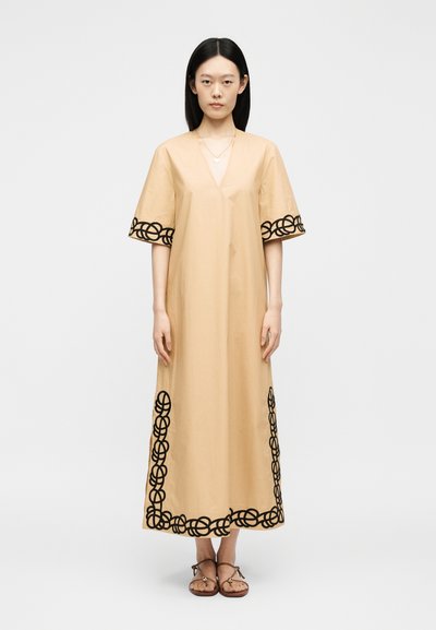 Femme aux longs cheveux noirs, portant une robe beige longueur cheville avec des motifs circulaires noirs sur les manches et l’ourlet, debout les bras le long du corps.