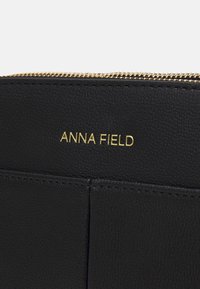 Czarny skórzany portfel z fakturowaną powierzchnią, złotym suwakiem i tłoczonym logo "ANNA FIELD" na przedniej stronie.