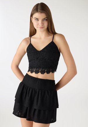 Giovane donna che indossa un top corto nero in pizzo floreale con spalline sottili e una gonna nera a strati, in piedi davanti a uno sfondo chiaro e uniforme.