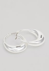 Pendientes de aro plateados con un acabado pulido y un logo de DKNY en el lado, exhibidos sobre un fondo neutro.
