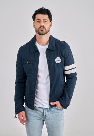 Donkerblauwe jas met rits, voorzien van een borstzak met knoop, gestreepte accenten op de mouwen en een logopatch aan de bovenkant van de linkerzijde.