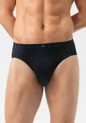 Männlicher Oberkörper, der schwarze Slips mit kleinen hellblauen Punkten vor einem einfarbigen hellen Hintergrund trägt.