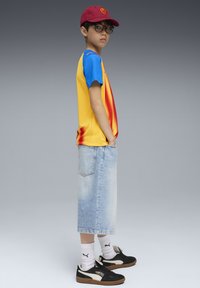 Ung person med röd keps, glasögon, gul och blå sporttröja, ljusblå denimkjol, vita Puma-strumpor och svarta Puma-sneakers som står i profil.