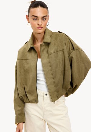 Mujer con el cabello recogido que lleva pendientes de aro de oro, una chaqueta oversize verde oliva, una camiseta de tirantes blanca con relieve y pantalones color crema.