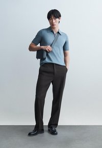 Mladi moški, ki nosi modro polo majico, črne hlače, črne čevlje in črno torbico čez ramo, stoji ob običajni sivi steni.