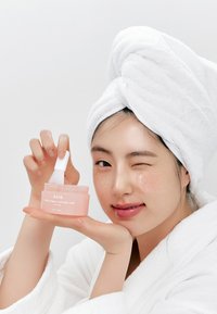 Maschera notturna al collagene rosa in un barattolo trasparente con coperchio bianco. Persona con un asciugamano bianco sulla testa tiene il barattolo, sorridendo.