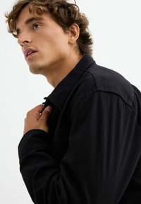 Camisa negra con un diseño de cuello, tela texturizada, costuras visibles y un solo bolsillo en el pecho, que es llevada por una persona que se ajusta el cuello.