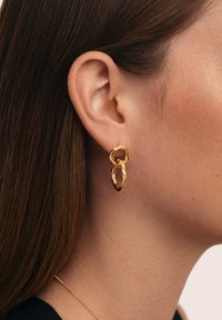 Pendientes de aro de oro con un diseño trenzado y texturizado que cuelga de un lóbulo perforado, presentando un aro más pequeño con cierre en la parte superior.