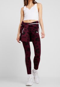 Bordowe legginsy z czarnym wzorem kwiatowym, białe paski po bokach, w połączeniu z białym prążkowanym crop topem i białymi trampkami.