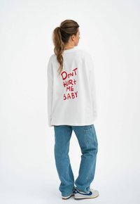 Weißes, übergroßes Sweatshirt mit rotem Text "DON'T HURT ME BABY" auf dem Rücken, kombiniert mit hellblauen Jeans und grauen Sneakern.