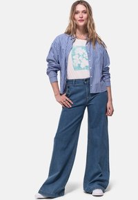 Camisa azul de rayas con botones, camiseta gráfica blanca con flores, jeans de denim de tiro alto y pierna ancha con botones laterales, usados con zapatillas blancas.