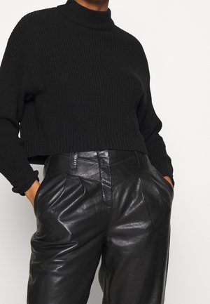 Femme portant un pull col roulé noir côtelé rentré dans un pantalon en cuir noir taille haute, les mains dans les poches.