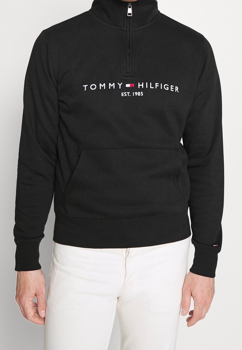 Homme portant un sweat-shirt noir à demi-fermeture éclair avec le logo "Tommy Hilfiger Est. 1985" et un pantalon blanc sur fond uni.