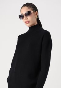 Fekete garbópulóver bordázott nyakkal. A modellen oversized, téglalap alakú napszemüveg van, sötét lencsékkel és arany részletekkel a kereten.