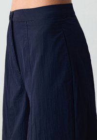 Donkerblauwe broek van een lichtgewicht, gestructureerde stof. Voorzien van een gladde tailleband, zijplooien en strakke lijnen voor een gepolijste uitstraling.