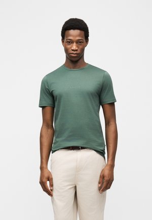 Mladi moški s kratkimi kodrastimi lasmi, oblečen v zeleno majico znamke Guess in svetlo bež hlače, stoji pred enostavnim svetlim ozadjem.