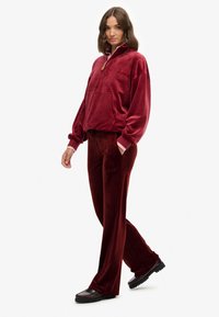 Kvinde iført matchende bordeaux velour zip-up trøje og bukser, stående med den ene hånd i lommen og iført mørke loafers.