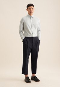 Camicia a righe verde chiaro e bianca, abbinata a pantaloni a pieghe blu navy e scarpe nere con lacci. Tessuto liscio e vestibilità su misura.