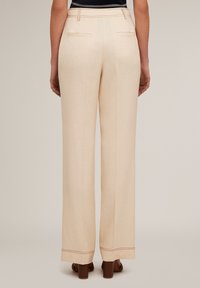 Pantalons beige à large jambe en tissu lisse, dotés de poches arrière, de coutures décoratives et d'une longueur raccourcie au-dessus de chaussures à talons beiges.