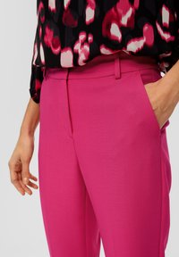 Strakke roze broek met zijzakken en vlakke voorkant. De stof lijkt glad en lichtjes gestructureerd. De hand van het model rust op de tailleband.